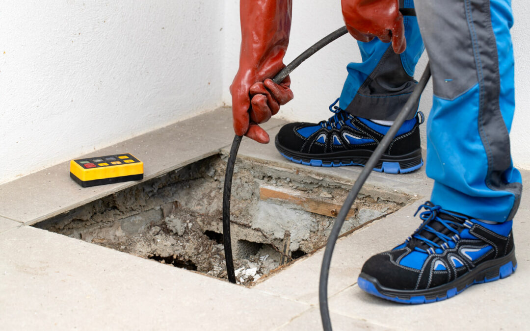 Worker in safety gear using a cable tool to inspect or clear a drain through a hole in a tiled floor.