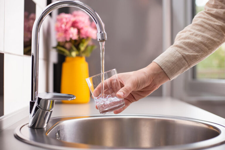 Hand filling a glass with clean water from a kitchen faucet, symbolizing safe drinking water ensured by backflow prevention.