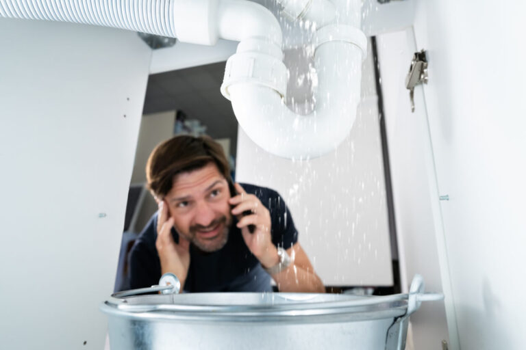 Frustrated man on the phone calling a plumber as water leaks heavily from a broken pipe under the sink into a bucket.