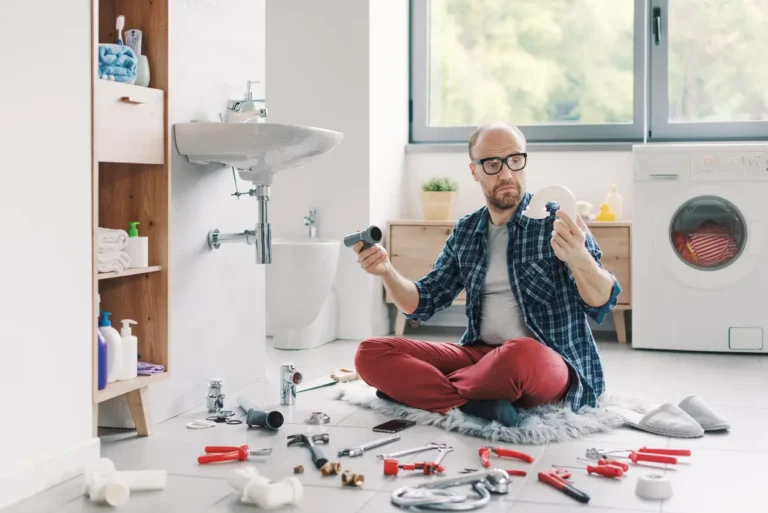 Man on bathroom floor surrounded by tools working with pipes, preparing for plumbing fixture installation under the sink.